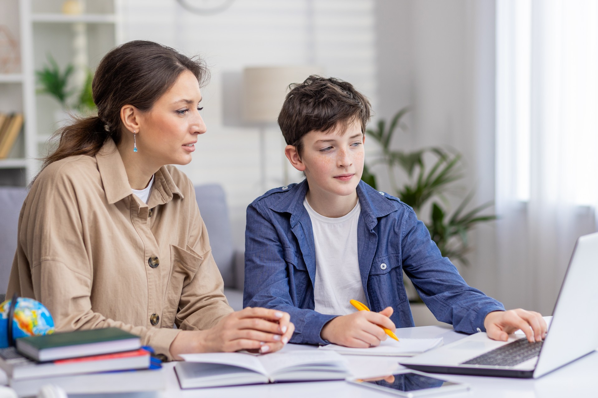 Mother and son studying together sitting at desk at home in living room, boy and woman tutor teaching at home, doing homework together